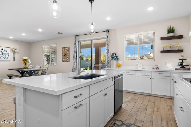 kitchen featuring stainless steel dishwasher, a sink, light countertops, light wood-type flooring, and a kitchen island with sink