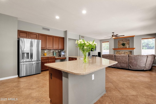 kitchen with stainless steel fridge with ice dispenser, a center island with sink, ceiling fan, backsplash, and a large fireplace