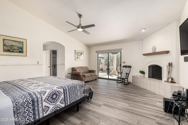 bedroom with arched walkways, a tiled fireplace, lofted ceiling, wood finished floors, and access to outside