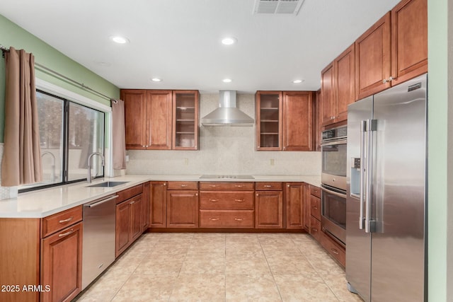 kitchen featuring a sink, light countertops, appliances with stainless steel finishes, wall chimney exhaust hood, and glass insert cabinets