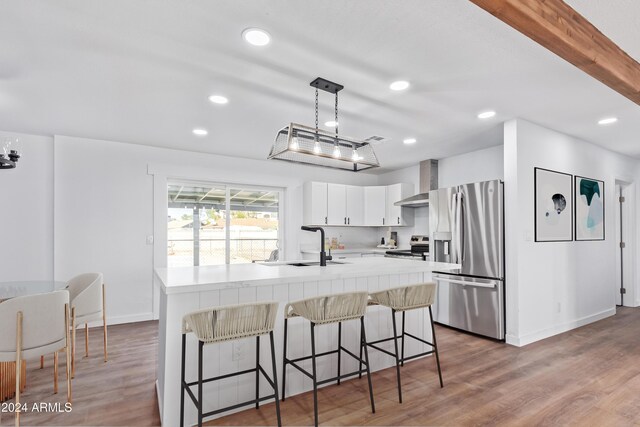 kitchen featuring wall chimney exhaust hood, stainless steel appliances, white cabinets, hanging light fixtures, and a breakfast bar area