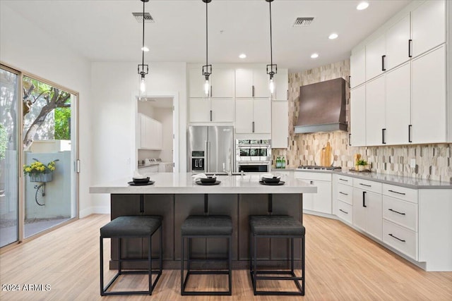 kitchen featuring light hardwood / wood-style flooring, appliances with stainless steel finishes, premium range hood, an island with sink, and white cabinetry