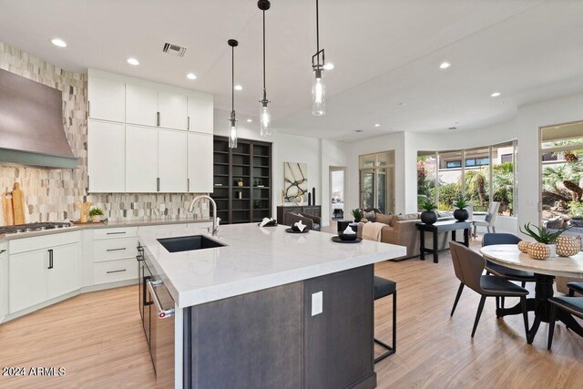 kitchen with appliances with stainless steel finishes, premium range hood, light hardwood / wood-style floors, an island with sink, and hanging light fixtures