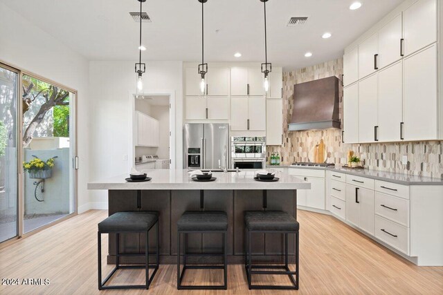 kitchen featuring premium range hood, light stone countertops, an island with sink, sink, and white cabinets