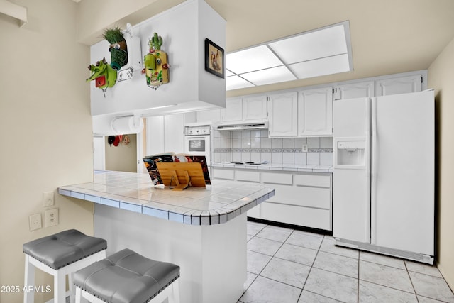 kitchen featuring tile counters, tasteful backsplash, a peninsula, white appliances, and under cabinet range hood