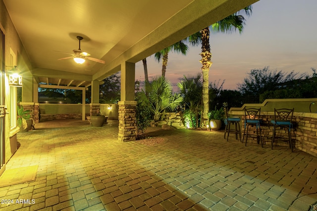 patio terrace at dusk with ceiling fan, exterior kitchen, and an outdoor bar