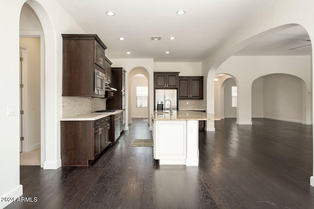 kitchen with ceiling fan, an island with sink, tasteful backsplash, dark brown cabinets, and dark hardwood / wood-style floors