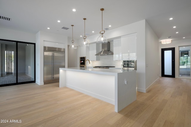 kitchen with wall chimney range hood, stainless steel appliances, backsplash, white cabinetry, and light wood-type flooring