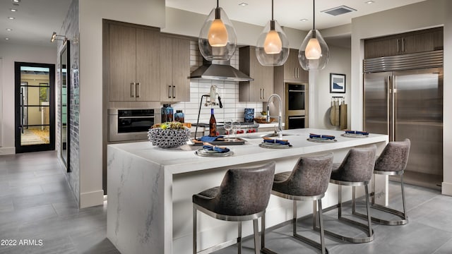 kitchen with tasteful backsplash, pendant lighting, wall chimney exhaust hood, a kitchen island with sink, and appliances with stainless steel finishes