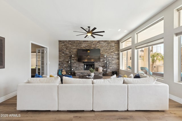 living area featuring ceiling fan, a stone fireplace, wood finished floors, and baseboards