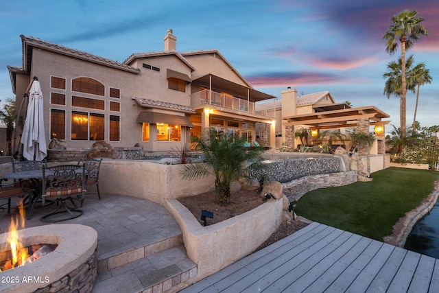 back of house at dusk with a patio, a balcony, outdoor dining space, a chimney, and stucco siding