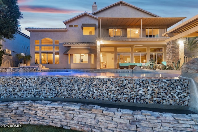 back of house at dusk with a chimney, stucco siding, a balcony, an outdoor pool, and a tiled roof