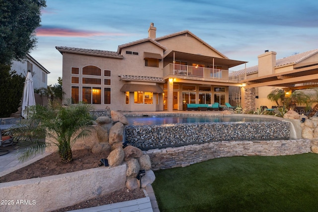 back of house at dusk with a tile roof, a chimney, stucco siding, a balcony, and an outdoor pool