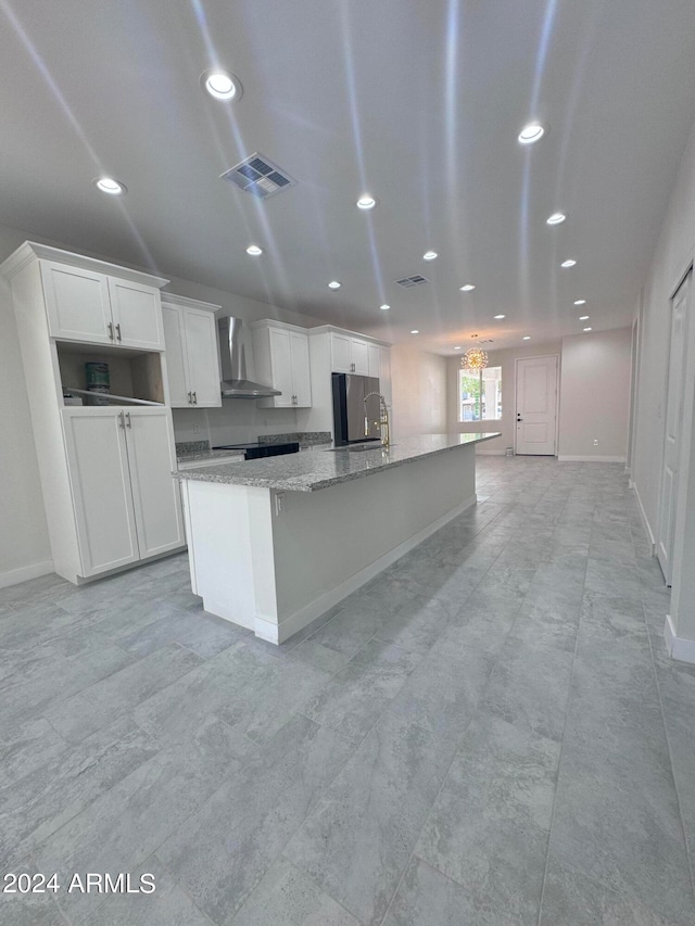 kitchen featuring stainless steel refrigerator, light stone counters, an island with sink, white cabinets, and wall chimney range hood