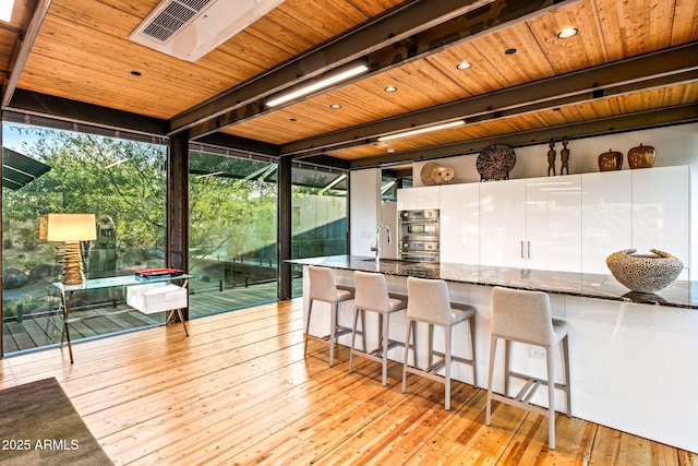 kitchen featuring wooden ceiling, light wood-type flooring, floor to ceiling windows, dark stone counters, and a kitchen bar