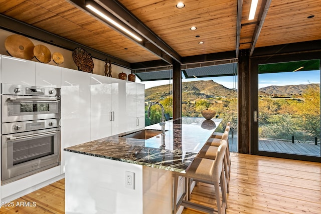 kitchen featuring double oven, wooden ceiling, a sink, light wood-type flooring, and dark stone countertops