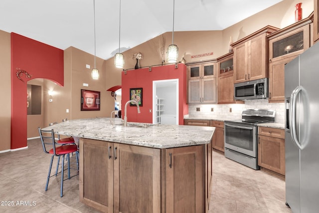 kitchen with brown cabinetry, decorative backsplash, appliances with stainless steel finishes, arched walkways, and a kitchen island with sink