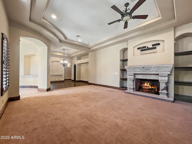 unfurnished living room with a warm lit fireplace, a raised ceiling, carpet floors, ceiling fan with notable chandelier, and baseboards