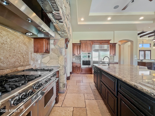 kitchen with wall chimney range hood, high quality appliances, arched walkways, stone tile floors, and decorative backsplash