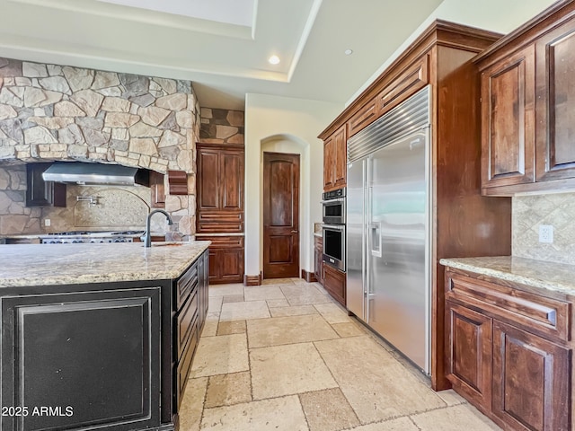 kitchen featuring light stone countertops, stainless steel built in fridge, backsplash, under cabinet range hood, and stone tile flooring