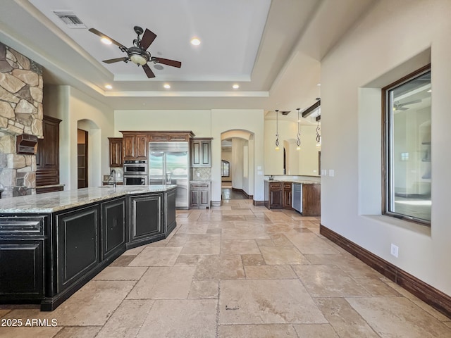 kitchen featuring a raised ceiling, arched walkways, stainless steel appliances, a ceiling fan, and stone tile flooring