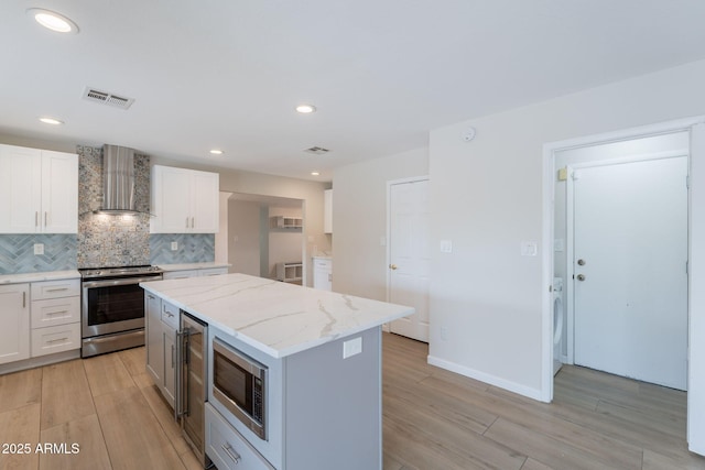 kitchen featuring stainless steel appliances, wall chimney range hood, light wood-type flooring, a center island, and decorative backsplash