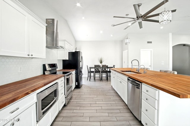 kitchen with wall chimney exhaust hood, a ceiling fan, a sink, stainless steel appliances, and wooden counters