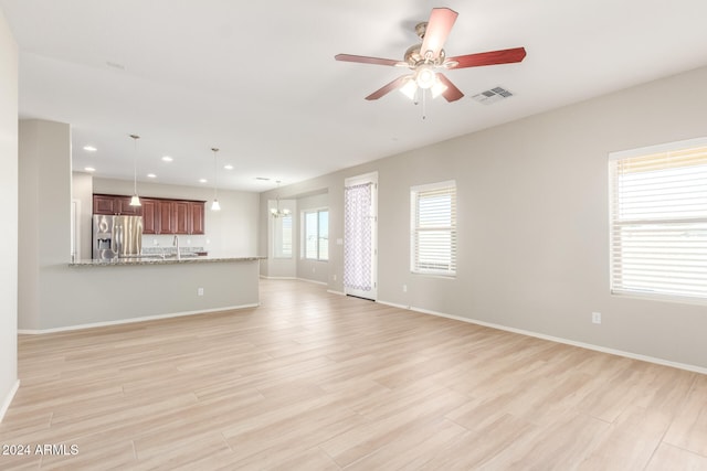 unfurnished living room featuring ceiling fan with notable chandelier, light wood-type flooring, and plenty of natural light