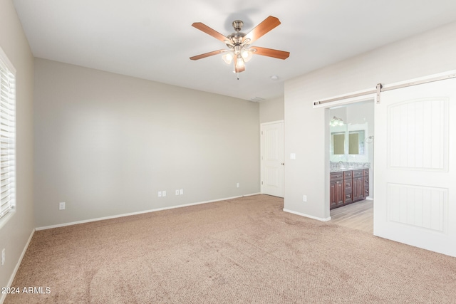 unfurnished bedroom with ensuite bathroom, a barn door, light carpet, and ceiling fan