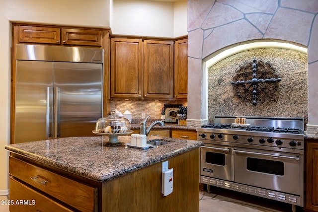 kitchen with high quality appliances, backsplash, dark stone counters, and an island with sink