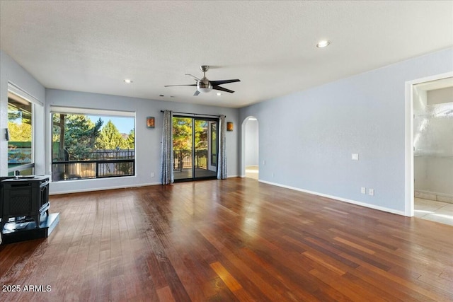 unfurnished living room with a ceiling fan, arched walkways, a wood stove, and wood finished floors