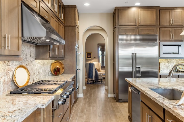 kitchen featuring light wood finished floors, under cabinet range hood, light stone counters, stainless steel appliances, and a sink