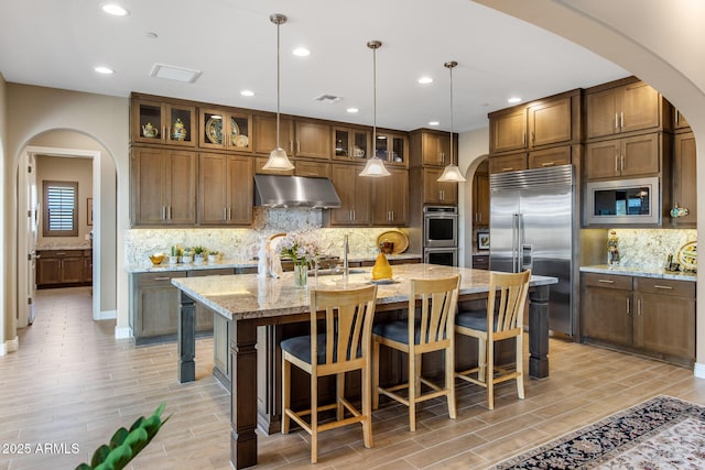 kitchen featuring a kitchen island with sink, under cabinet range hood, arched walkways, built in appliances, and wood tiled floor