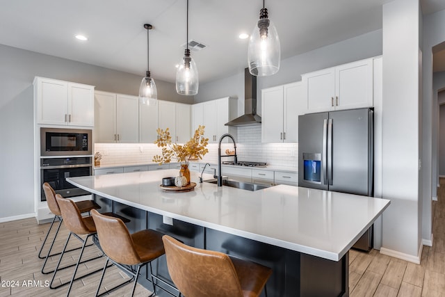 kitchen featuring stainless steel appliances, a center island with sink, wall chimney exhaust hood, a kitchen breakfast bar, and white cabinetry