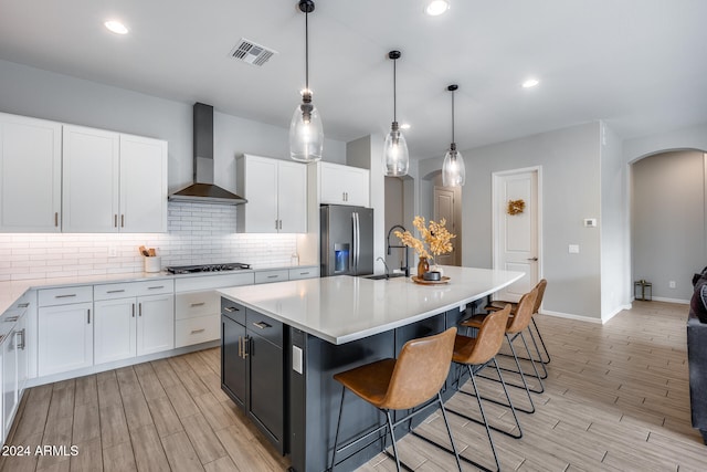 kitchen with white cabinetry, appliances with stainless steel finishes, hanging light fixtures, wall chimney exhaust hood, and a kitchen island with sink