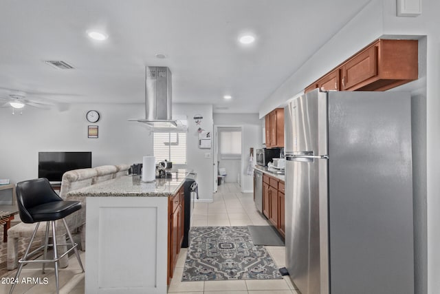 kitchen with ceiling fan, stainless steel appliances, light tile floors, a kitchen breakfast bar, and wall chimney exhaust hood