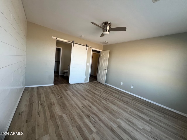 unfurnished bedroom with ensuite bathroom, a barn door, ceiling fan, and wood-type flooring