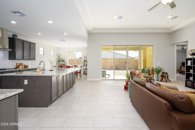 kitchen with dark brown cabinets, backsplash, light stone countertops, wall chimney exhaust hood, and sink