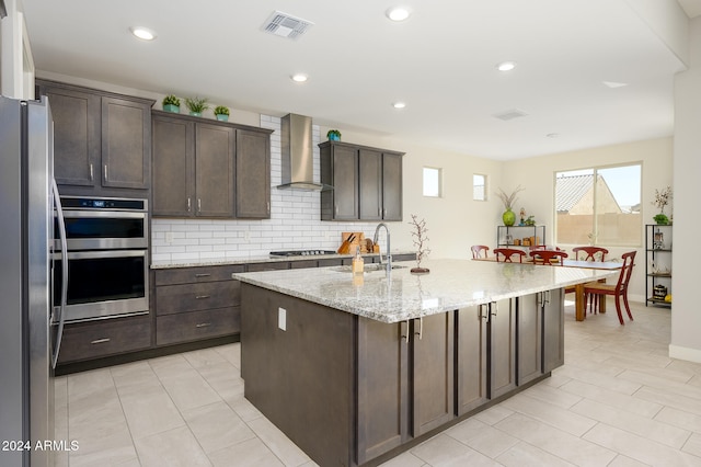 kitchen with wall chimney exhaust hood, an island with sink, stainless steel appliances, sink, and light stone counters