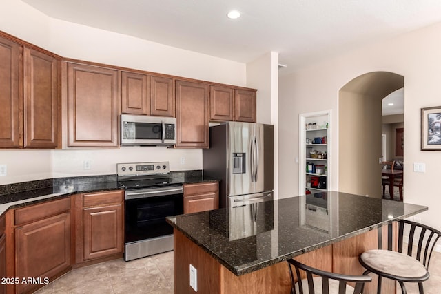 kitchen with arched walkways, brown cabinetry, dark stone counters, appliances with stainless steel finishes, and a breakfast bar area