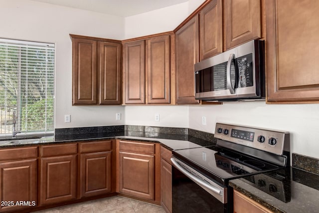 kitchen with light tile patterned floors, stainless steel appliances, a sink, dark stone counters, and brown cabinetry