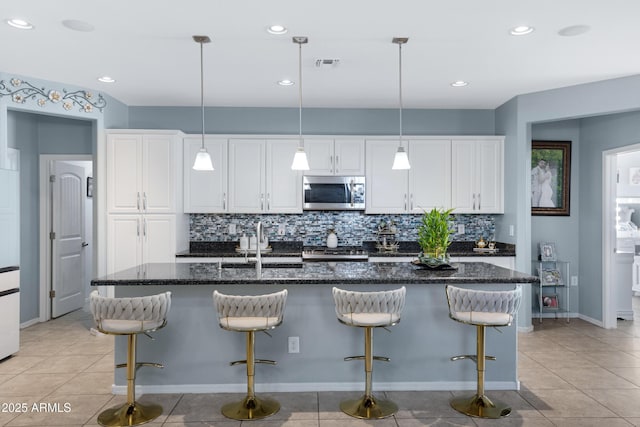 kitchen with an island with sink, light tile patterned floors, white cabinets, and dark stone counters