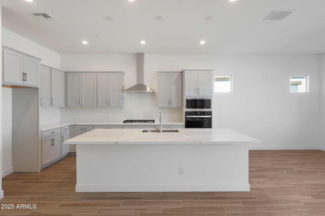 kitchen featuring light stone counters, gray cabinets, a kitchen island with sink, wall chimney range hood, and black oven