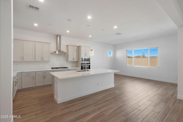 kitchen featuring wall chimney exhaust hood, a kitchen island with sink, light stone counters, gas cooktop, and stainless steel microwave