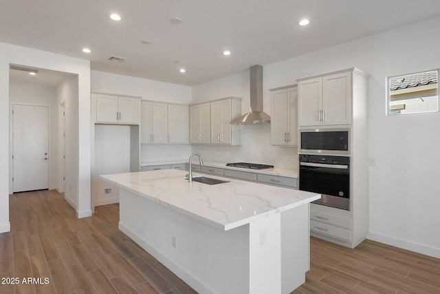 kitchen featuring sink, wall chimney range hood, stainless steel appliances, and a kitchen island with sink