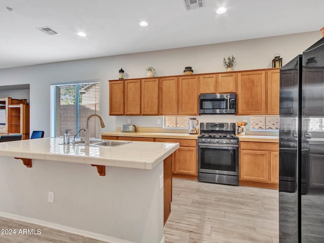 kitchen featuring sink, tile countertops, a kitchen island with sink, appliances with stainless steel finishes, and light wood-type flooring