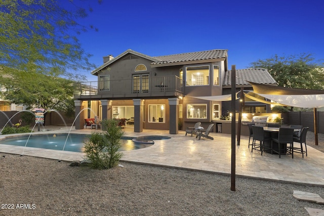 back house at dusk featuring a patio area, a pool with hot tub, pool water feature, a balcony, and an outdoor kitchen