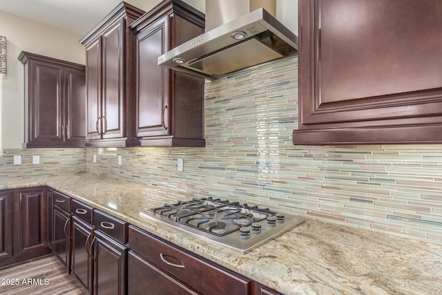 kitchen with stainless steel gas cooktop, decorative backsplash, dark brown cabinets, light stone countertops, and wall chimney exhaust hood