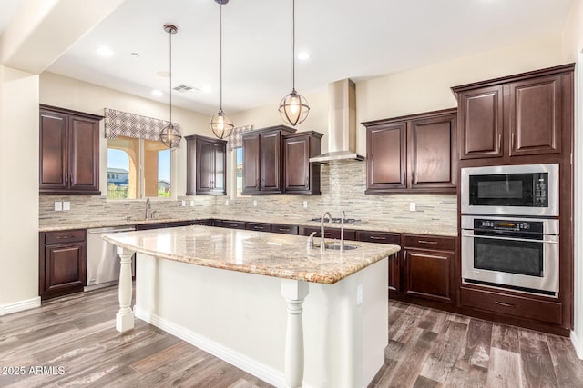 kitchen featuring a center island with sink, appliances with stainless steel finishes, a sink, wall chimney range hood, and wood finished floors