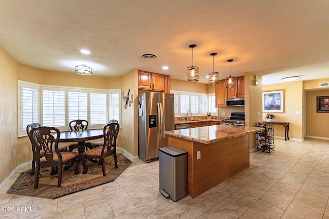 kitchen featuring visible vents, brown cabinetry, a kitchen island, appliances with stainless steel finishes, and light stone counters